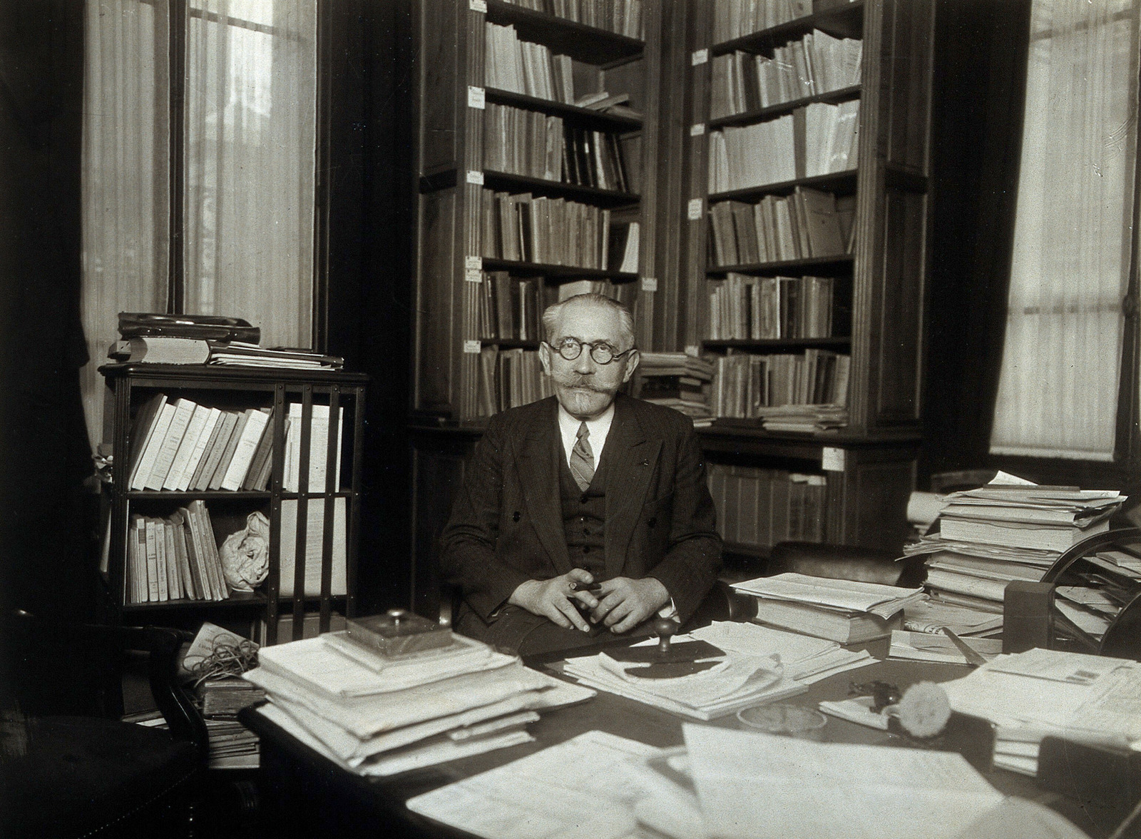 Paul Langevin sits behind a desk covered with papers, surrounded by bookshelves.